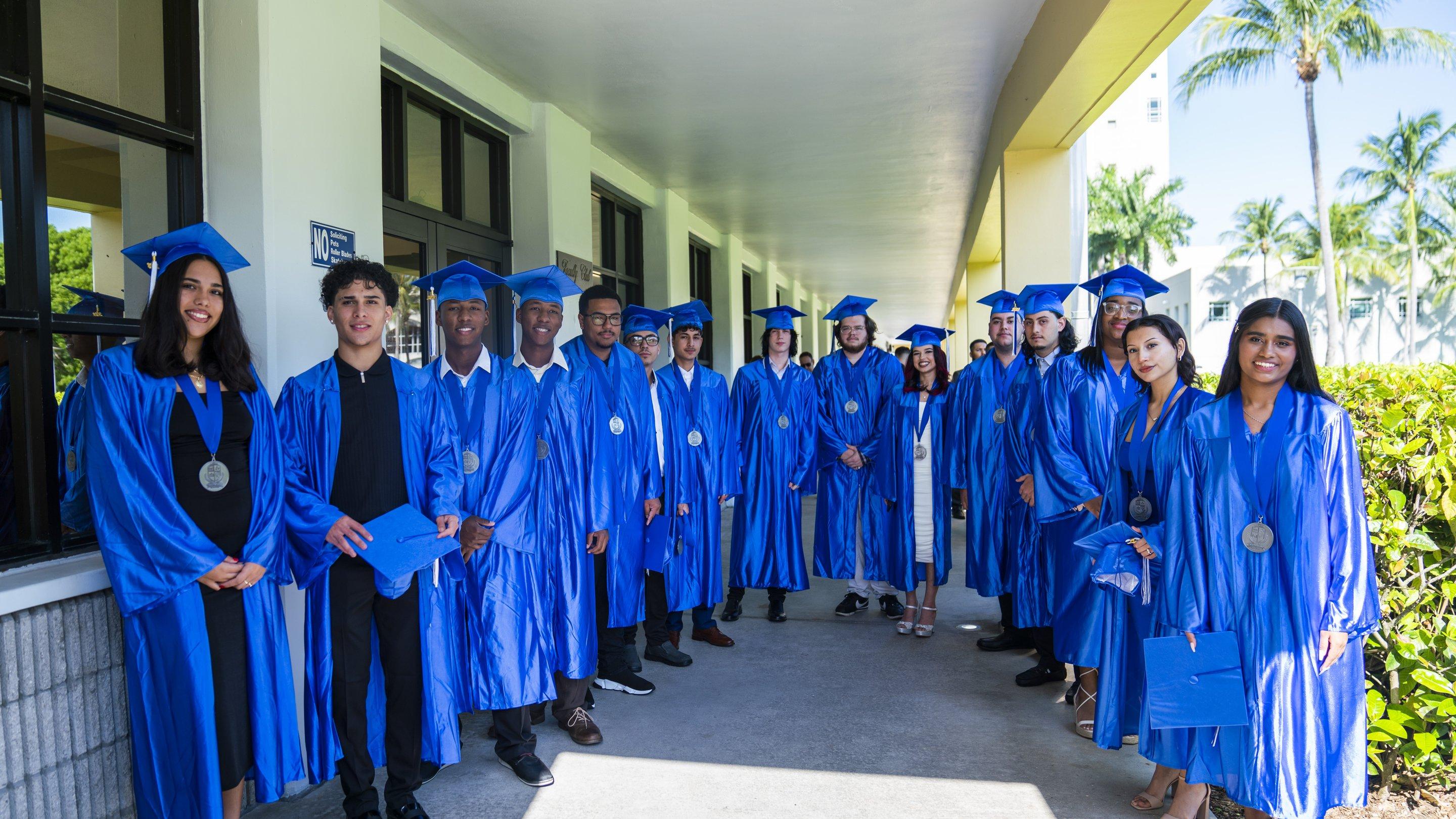 A line of graduates in blue caps and gowns outdoors under palm trees